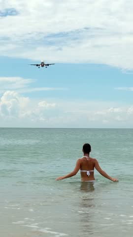 Vertical video. A young woman swims in the ocean on a sunny day when a plane flies overhead. Travel, tourism and air flights concept
