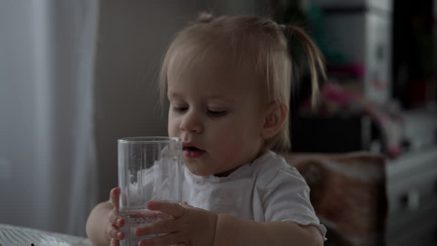 Charming little girl with two ponytails sitting at dining table, drinking fresh water from transparent glass, with plate of almost finished meal in front of her, enjoying lunch break, slow motion.