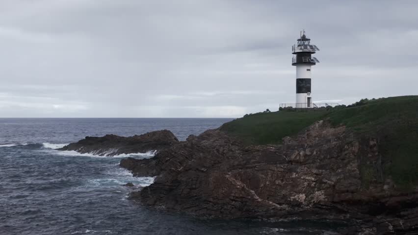 Video of the Illa Pancha lighthouse in Galicia, Spain, filmed on a cloudy day. The sea crashes against the rugged rocks of the island, creating a dramatic and atmospheric coastal scene.