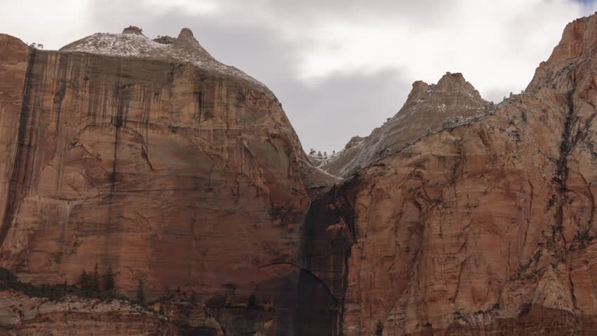 A wind whipped ephemeral waterfall pours over the cliff between The Streaked Wall and The Sentinel in Zion NP Ut. USA while dark clouds stream overhead and snow begins to fall blowing into the camera.