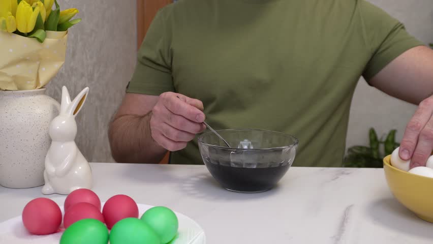 A man paints Easter eggs at home, showing the process of lowering a white egg into a bowl with a purple coloring solution using a spoon