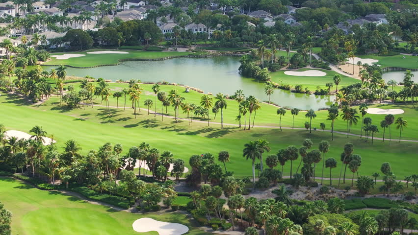An aerial view showcases Floridas beautiful and vibrant green golf courses, highlighting stunning greens, peaceful water bodies, and elegant palm trees all under a clear, sunny sky