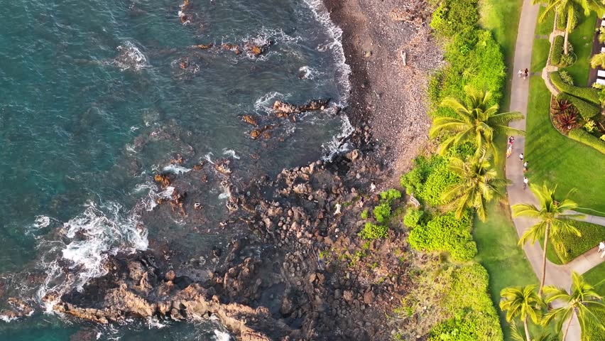 Aerial view of beautiful turquiose sea waves breaking on lava rock coastline with people walking along beach path and green grass shoreline in Wailea Maui, Hawaii during sunset landscape - 4K Drone