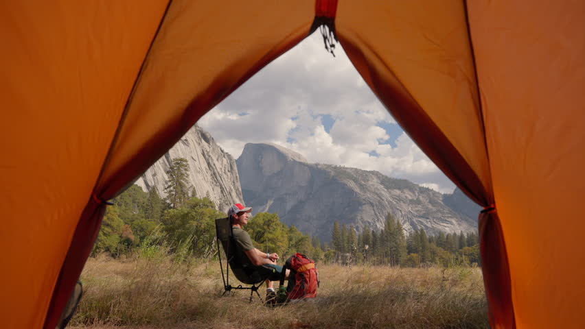 A serene view from an orange tent in Yosemite National Park, featuring a man relaxing amidst stunning landscapes, with El Capitan and Half Dome majestically rising in the background