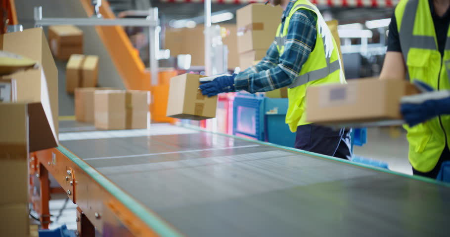 Efficient Logistics Team in Safety Vests Sorting and Loading Packages on a Conveyor Belt, Working in a Busy Distribution Center. Young Male and Female Handling Packages for Outgoing E-Commerce Orders