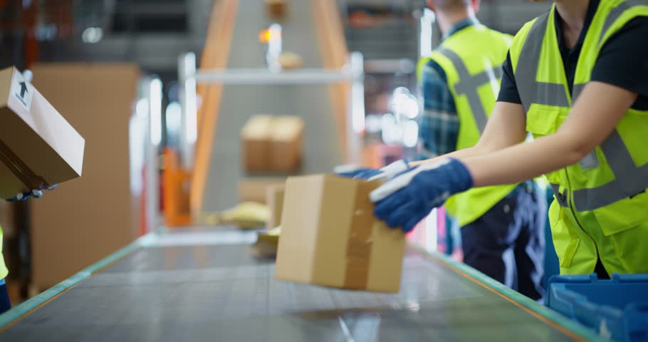 Efficient Logistics Team in Safety Vests Sorting and Loading Packages on a Conveyor Belt, Working in a Busy Distribution Center. Young Male and Female Handling Packages for Outgoing E-Commerce Orders