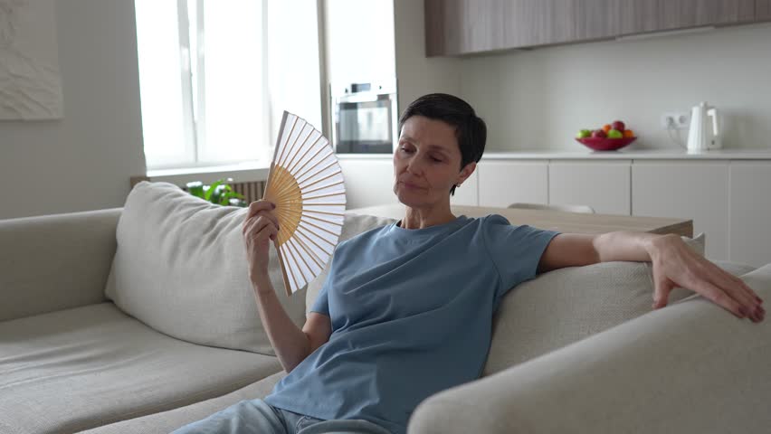 Unwell exhausted mature woman sitting on home couch waving paper fan to cool down. Overheated female suffers from stuffiness, feeling hot during heatwave in apartment without air conditioning. 