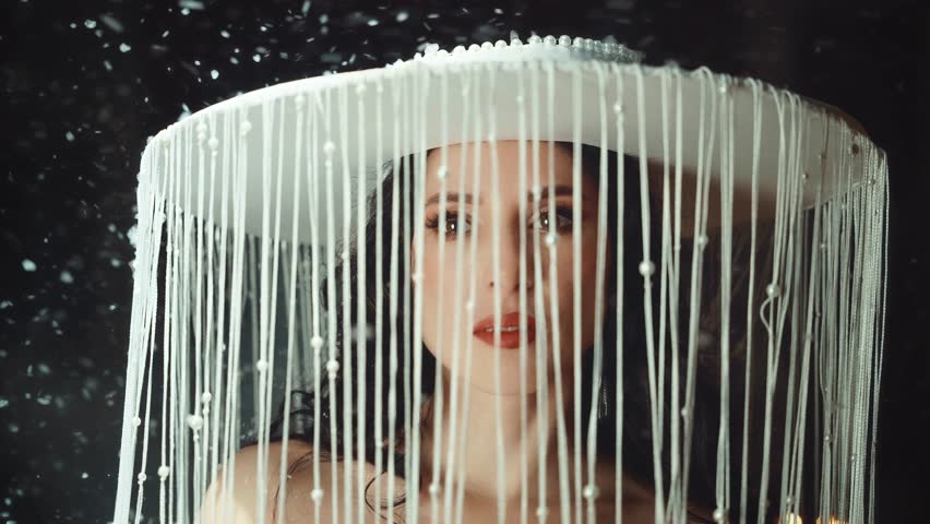 Fantasy woman queen in white wide-brimmed hat on head with strings of pearls hide face, sexy eyes looking at camera, fairy tale girl under falling snow night winter nature. magic New Year holiday