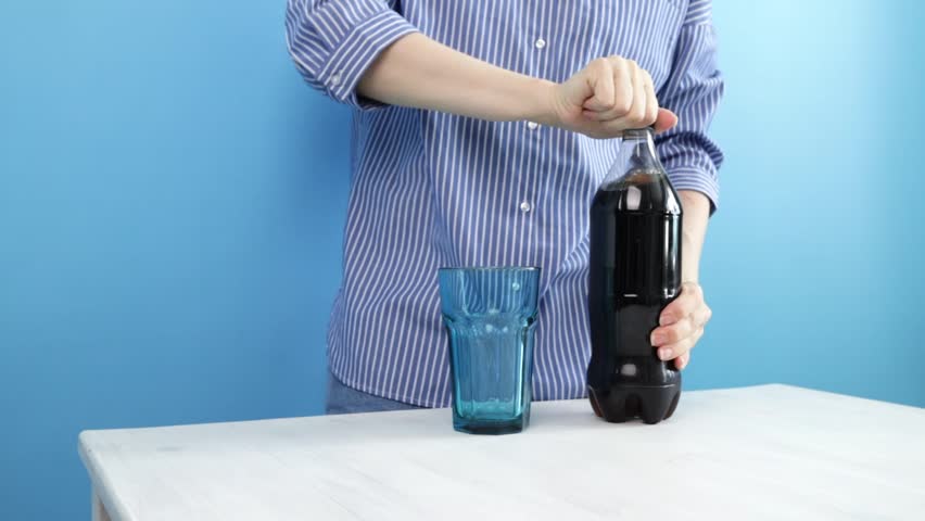 Female hands pouring soda cola into a glass from a bottle, refreshing beverage in a transparent glass, carbonated liquid for hydration on a blue background.