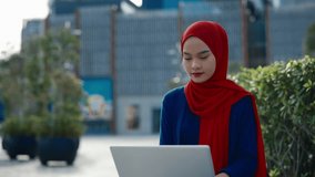 Portrait of young Muslim woman office employee typing on laptop in urban park. Malaysian female corporate professional works online against downtown buildings. Business career - Powered by Shutterstock - Get 15% off with code: PIKWIZARD15