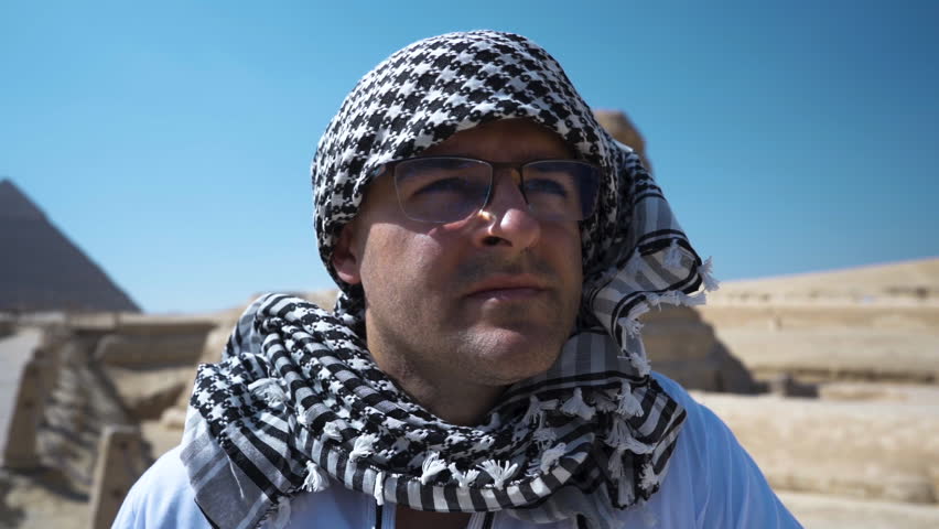 Portrait of a Bedouin man in glasses looking into the distance at the horizon, a sphinx in the background. An Egyptian man contemplates what is happening in the distance