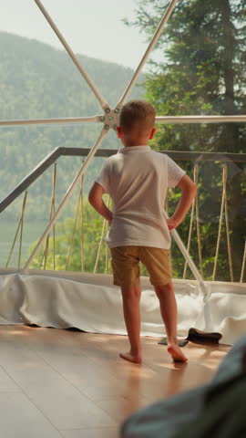 Child exercises by panoramic window at glamping. Little boy does yoga standing in clear camp cabin. Cute kid sports activity on family vacation