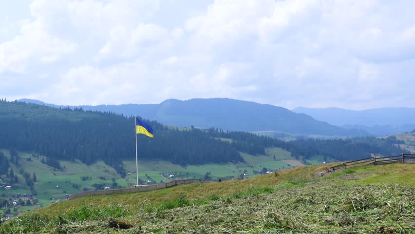Ukrainian flag flutters against the backdrop of a beautiful summer landscape of the Carpathian Mountains and the village of Verkhovyna. Ukraine. Blue cloudy sky and green spruce forests.