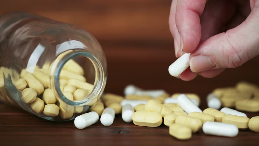 A hand showing white capsules and yellow pills next to a pill bottle. Nutritional dietary supplements, healthy lifestyle background.