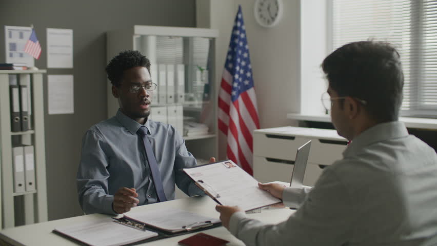 Black consular officer taking application form on clipboard and asking male applicant questions during visa interview at US embassy