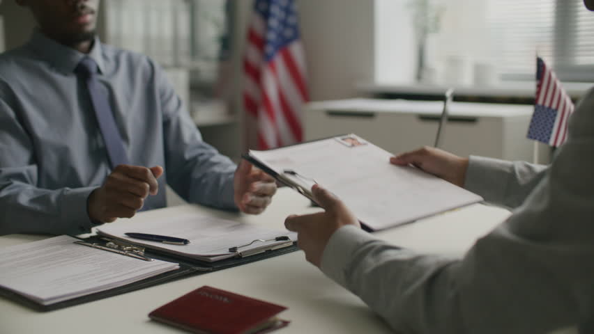 African American consular officer taking clipboard from applicant, reviewing documents and asking questions during visa interview at US embassy. Midsection, close-up shot