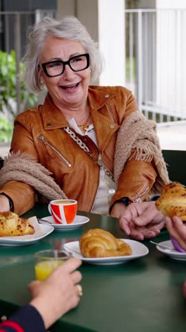 Vertical. Group laughing mature people having snack sitting at cafe bar. Older friends offering croissant gathered together in terrace. Food and beverage lifestyle in senior citizens concept