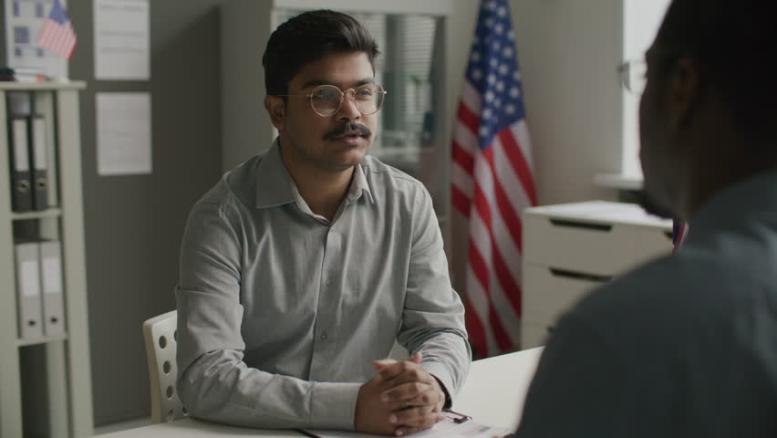Indian man sitting at office desk, talking to consul during visa interview in embassy with American flag in background. Over the shoulder view