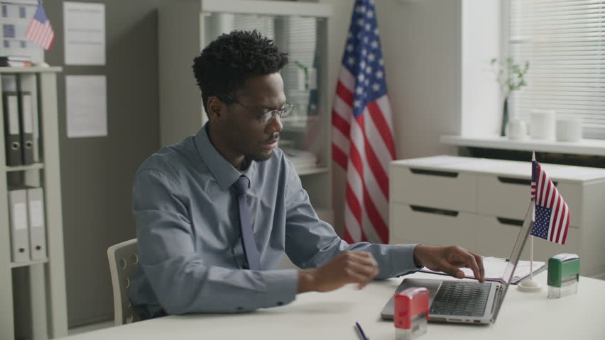 Black visa officer sitting at office desk, examining application form, stamping rejection on documents and typing on laptop during workday at American consulate