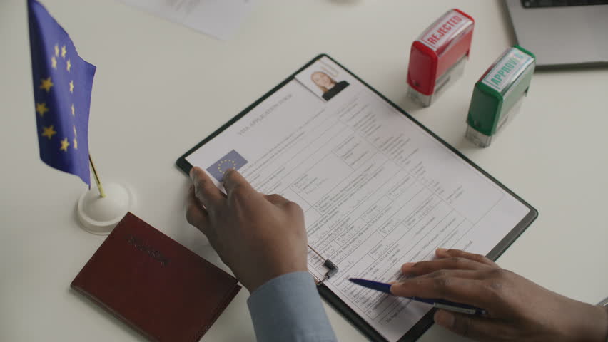 Hands of unrecognizable consular officer stamping APPROVED on European visa application form on clipboard with European Union flag and passport on desk nearby. Close-up view