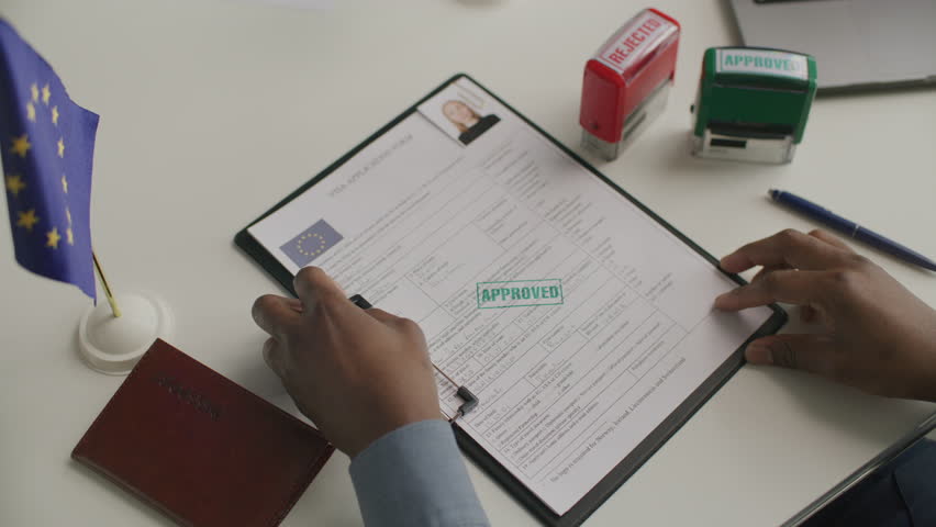 Hands of unrecognizable consular officer stamping APPROVED on European visa application form on clipboard with European Union flag and passport on desk nearby. Close-up view