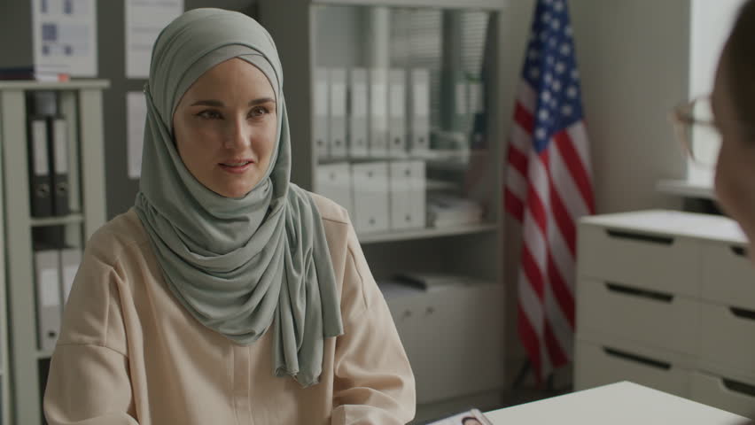 Female consular officer holding visa application form and speaking with young muslim woman in hijab during interview at US embassy. Tilt-up, over the shoulder view