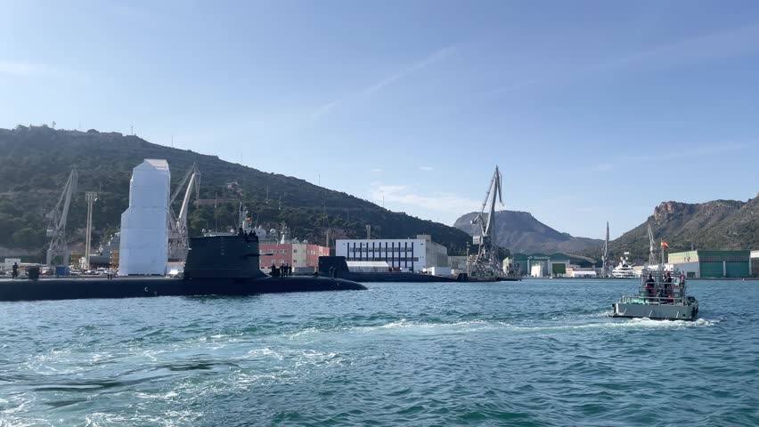 Submarine docked at naval base with boats and cranes in coastal area
