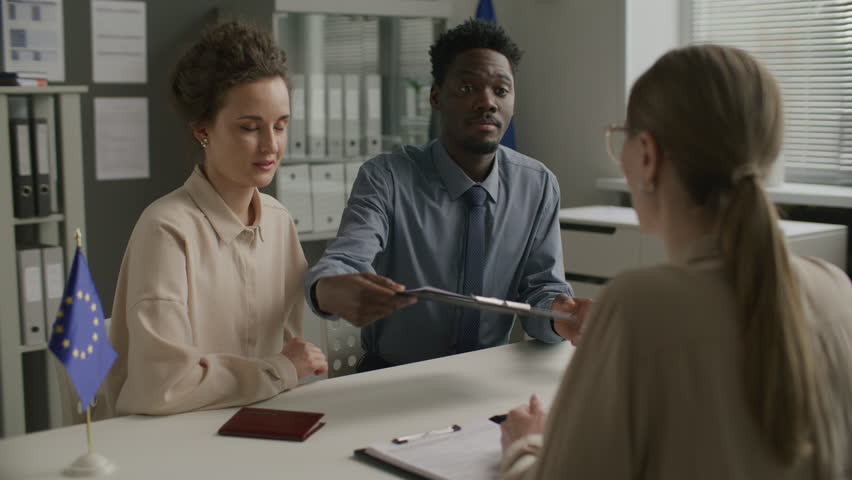 Young diverse couple smiling and answering questions from female consular officer while attending immigration interview at European Union embassy