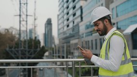Portrait of focused inspector in safety vest with smartphone walking along overpass bridge. Bearded man contractor in helmet scrolls social media via cellular at construction site - Powered by Shutterstock - Get 15% off with code: PIKWIZARD15