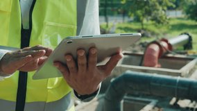 Engineer in safety vest inputs information on tablet at waste water treatment facility closeup. Technician analyzes digital data of sewer filtration station standing against pipes - Powered by Shutterstock - Get 15% off with code: PIKWIZARD15