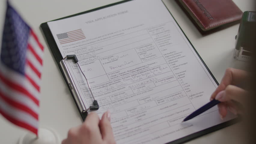 Hands of female consular officer examining and approving visa application form with official stamp at table with American flag and passport on it. Close-up view