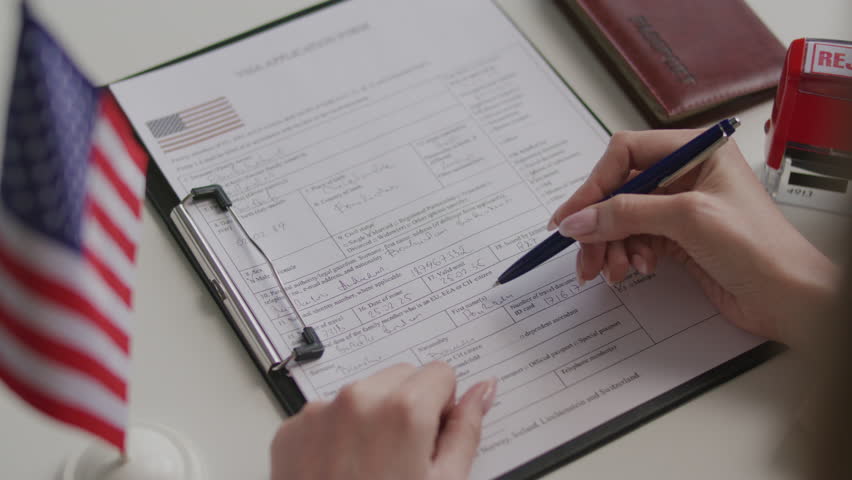 Hands of female consular officer stamping rejected mark on visa application form after reviewing it at desk with American flag and passport nearby. Close-up shot