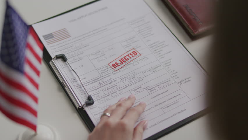 Hands of female consular officer stamping rejected mark on visa application form after reviewing it at desk with American flag and passport nearby. Close-up shot