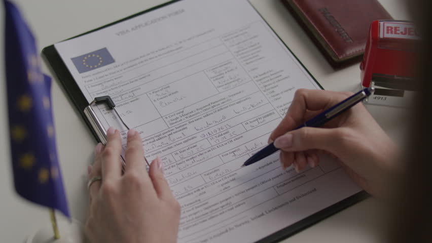 Hands of female consular officer pressing red rejected stamp on Schengen visa application form with EU flag and passport beside it on office desk. Close-up shot