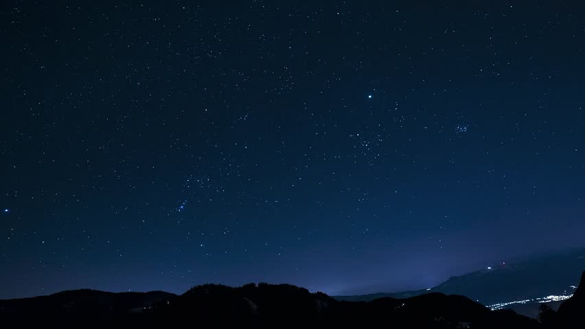 Timelapse showing stars moving across a clear night sky, with mountain silhouettes in the foreground and faint city lights at the base.