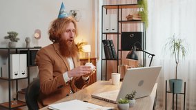 Happy Caucasian businessman wears festive birthday hat hold cupcake makes wish joyful congratulating blowing burning candle on cake. Redhead man freelancer celebrating birthday party at home office - Powered by Shutterstock - Get 15% off with code: PIKWIZARD15
