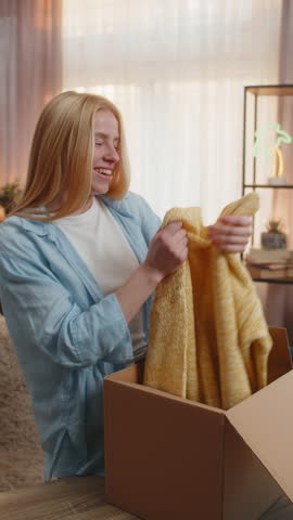 Young woman opens delivery box at home, removing taking blouse sweater clothing with excited grin and showing to camera. Caucasian girl feels satisfaction, enjoying new arrivals under evening sunset.