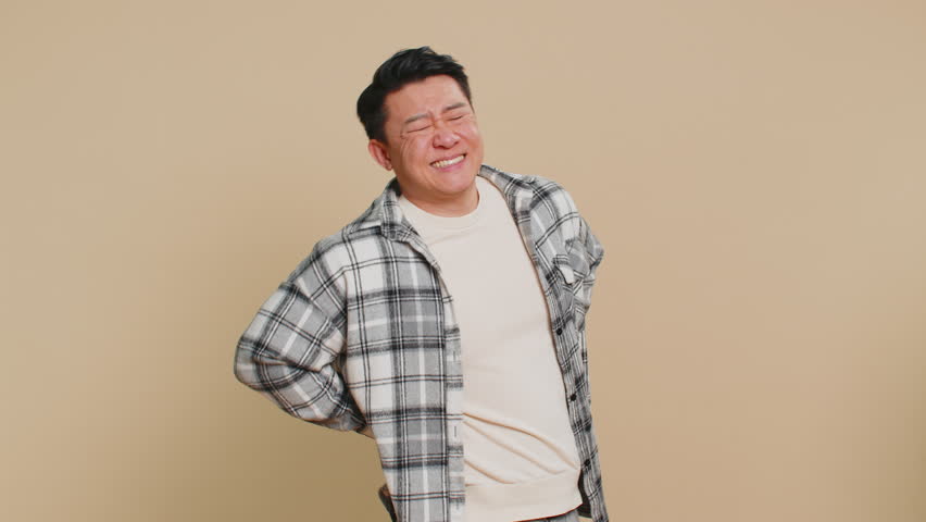 Asian middle-aged man suffering backache, massaging sore place lower spine, frowning from unbearable spasm torn muscles and tendons, kidney stones pinched nerve. Mature Chinese guy on beige background