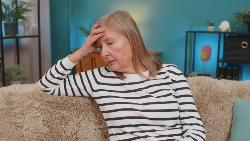 Caucasian mature woman places hand on forehead, sitting on sofa at home, showing frustration helplessness. Grandmother expresses stress disappointment after bad day, feeling overwhelmed disheartened