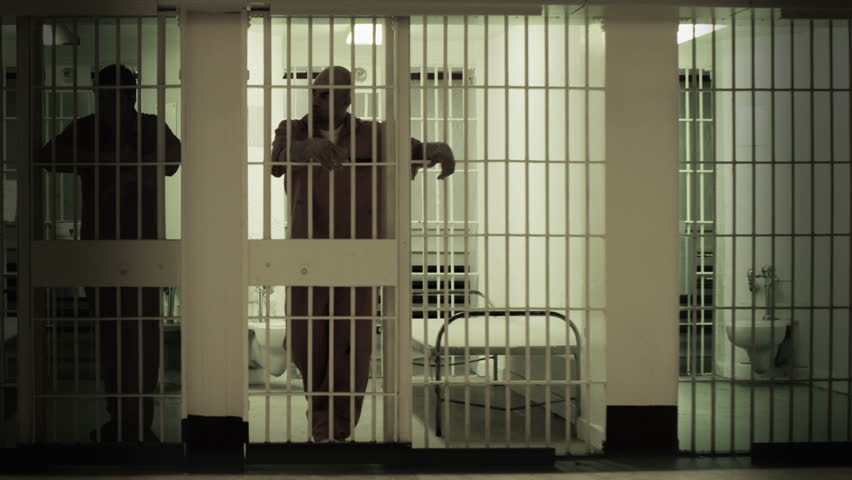 Inmate standing at the bars of his prison cell. Neighboring inmate can be seen in next cell. Desaturated color. Royalty-Free Stock Footage #3747254