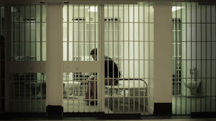 Inmate gets up from cot and stands at the bars in a prison cell. Desaturated