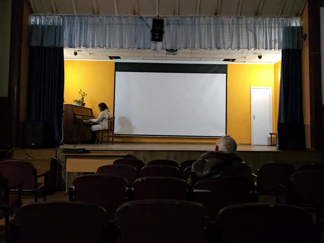 Man sits in a chair in front of a projector screen. A woman is playing the piano in the background in assembly hall