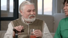 Portrait of positive handsome elderly grey haired bearded man holding walking stick, drinking fresh juice from glass and friendly chatting with black female nurse in long term care facilities. - Powered by Shutterstock - Get 15% off with code: PIKWIZARD15