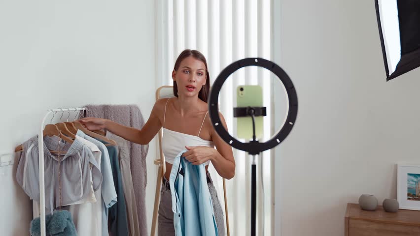 Young social medial content creator woman make fashion video. Blogger smiles to camera using selfie stick and light ring while making persuasive online clothing sell vlog to audience or follower. Adit