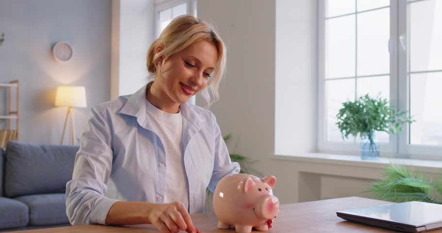 Happy woman putting a coin into a piggy bank at home, saving money for future investments. Finance, economy, and the importance of saving in a personal and joyful domestic setting.