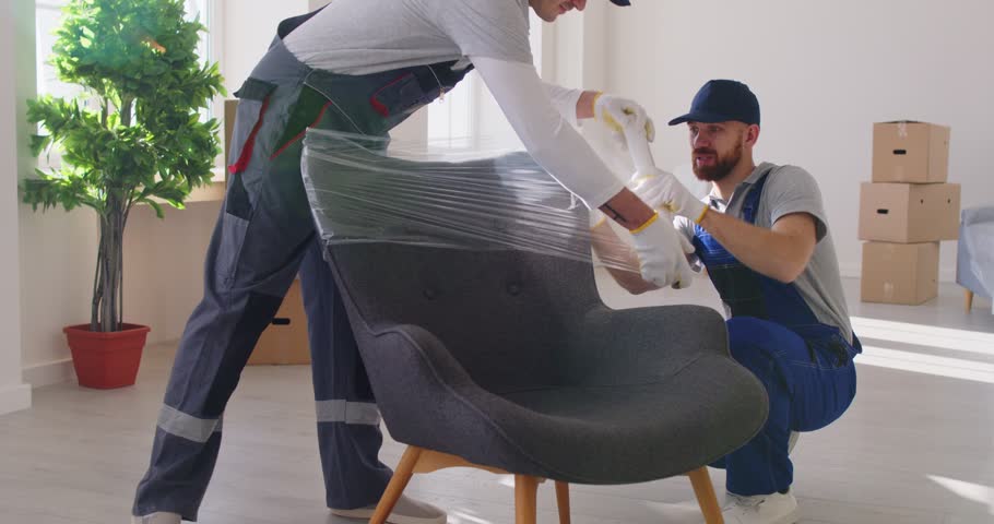 Portrait of a two young smiling male movers packing furniture in the living room. Moving service men workers wrapping chair with plastic wrap. Move, moving day and relocation concept. 4k video.