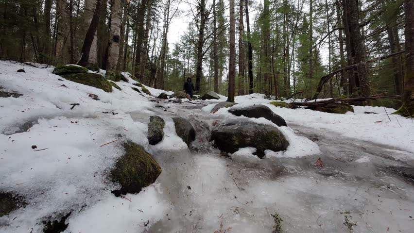A close up of water runoff as a Caucasian man hikes down a wet and slippery hill covered in melting snow and ice in Algonquin Park, Ontario, Canada