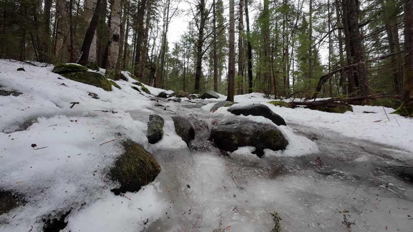 A close up of water runoff as a Caucasian man hikes up a wet and slippery hill covered in melting snow and ice in Algonquin Park, Ontario, Canada