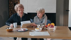 Two elderly women sitting at a wooden table, discussing a recipe book while preparing ingredients for cooking in a cozy home setting. High quality 4k footage - Powered by Shutterstock - Get 15% off with code: PIKWIZARD15