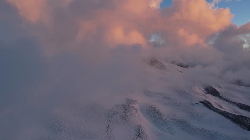 Aerial view of Mount Elbrus as the sun sets behind distant mountain peaks. The deep shadows and glowing sky create a dramatic contrast over the snow-covered landscape.